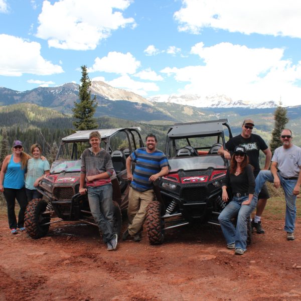 A group of people stand with two parked RZR ATV's with large mountains in the background
