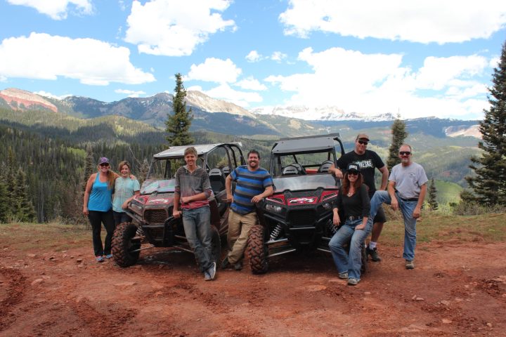 A group of people stand with two parked RZR ATV's with large mountains in the background