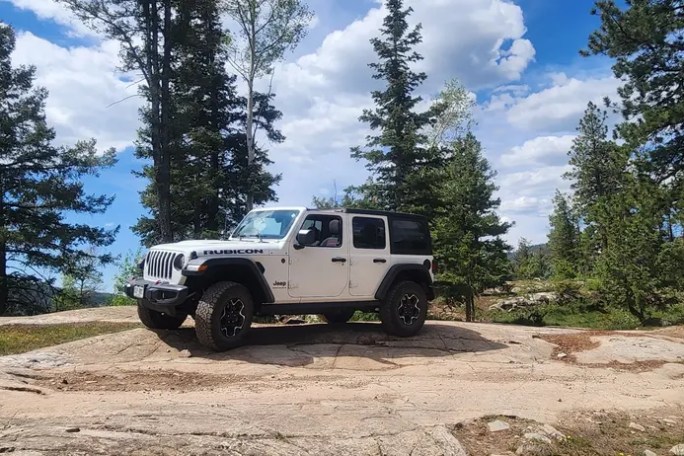 White Jeep parked on a rocky terrain surrounded by tall pine trees.